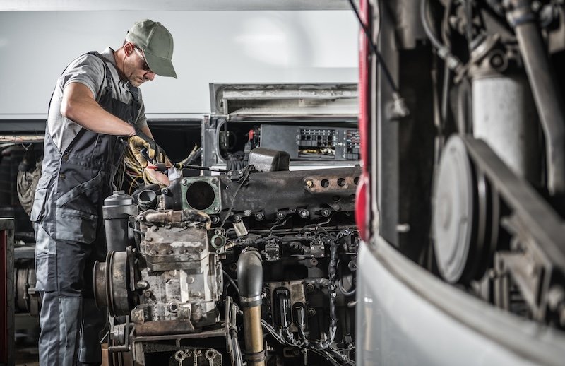 Professional Caucasian Automotive Mechanic in His 40s Repairing Powerful Heavy Duty Truck or Bus Diesel Engine Inside Truck Service Center. Transportation Industry Theme.