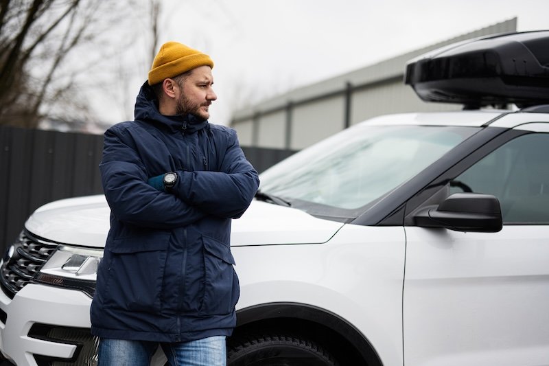 Man driver wear jacket and yellow hat against his american SUV car with roof rack in cold weather.